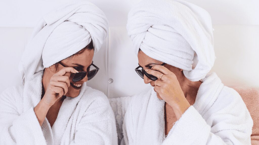 Two women enjoying a spa day, wearing towels and sunglasses, smiling and relaxing together.