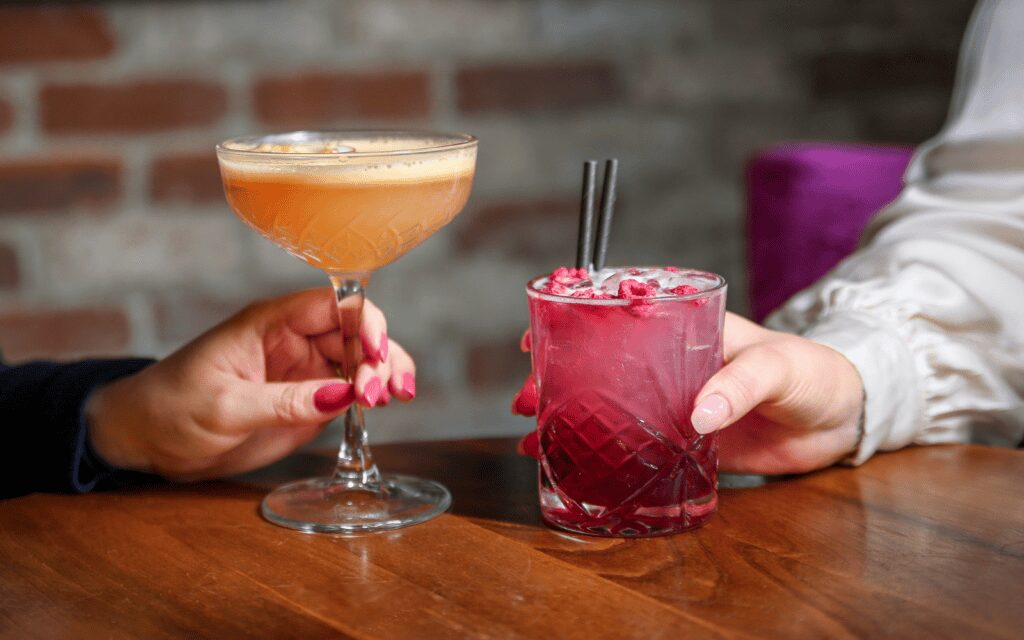 Two colourful cocktails, one pink and one orange, held by guests at a table.